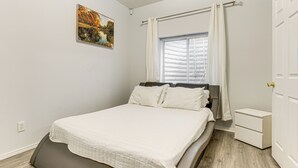 Un dormitorio con cama, mesita de noche, ventana con cortinas, puerta y un cuadro colgado en la pared.