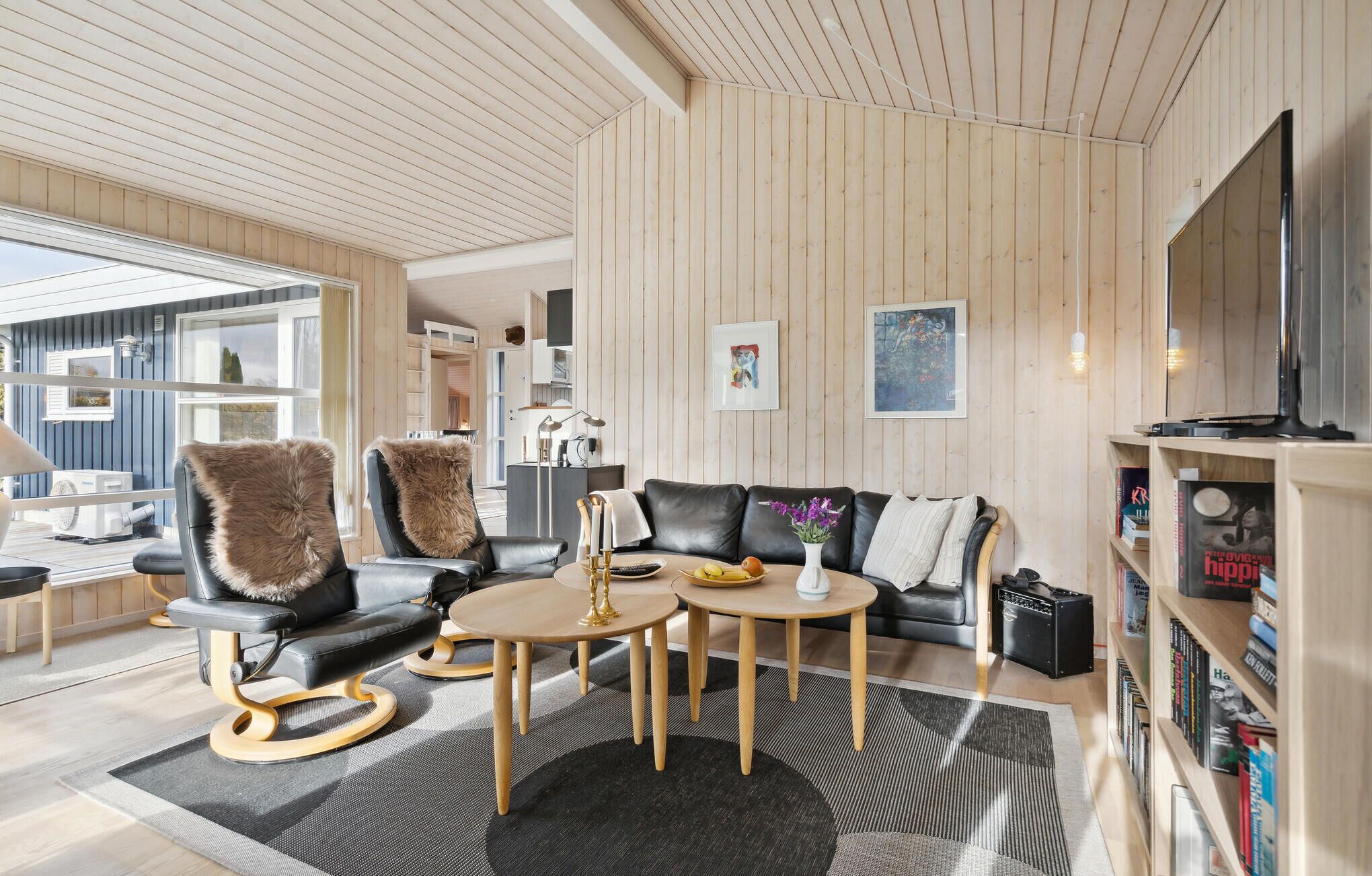 A modern living room with a black leather sofa, two armchairs with fur throws, a coffee table, and a bookshelf.