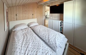 A bedroom with a wooden ceiling, a bed with patterned bedding, a white wardrobe, and a bookshelf.