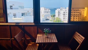 Un balcón con vista al mar y edificios, una pequeña mesa con una planta y asientos de madera.
