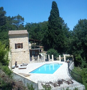 Un bâtiment en pierre doté d’une piscine, entouré d’arbres, avec un grand arbre visible au loin.