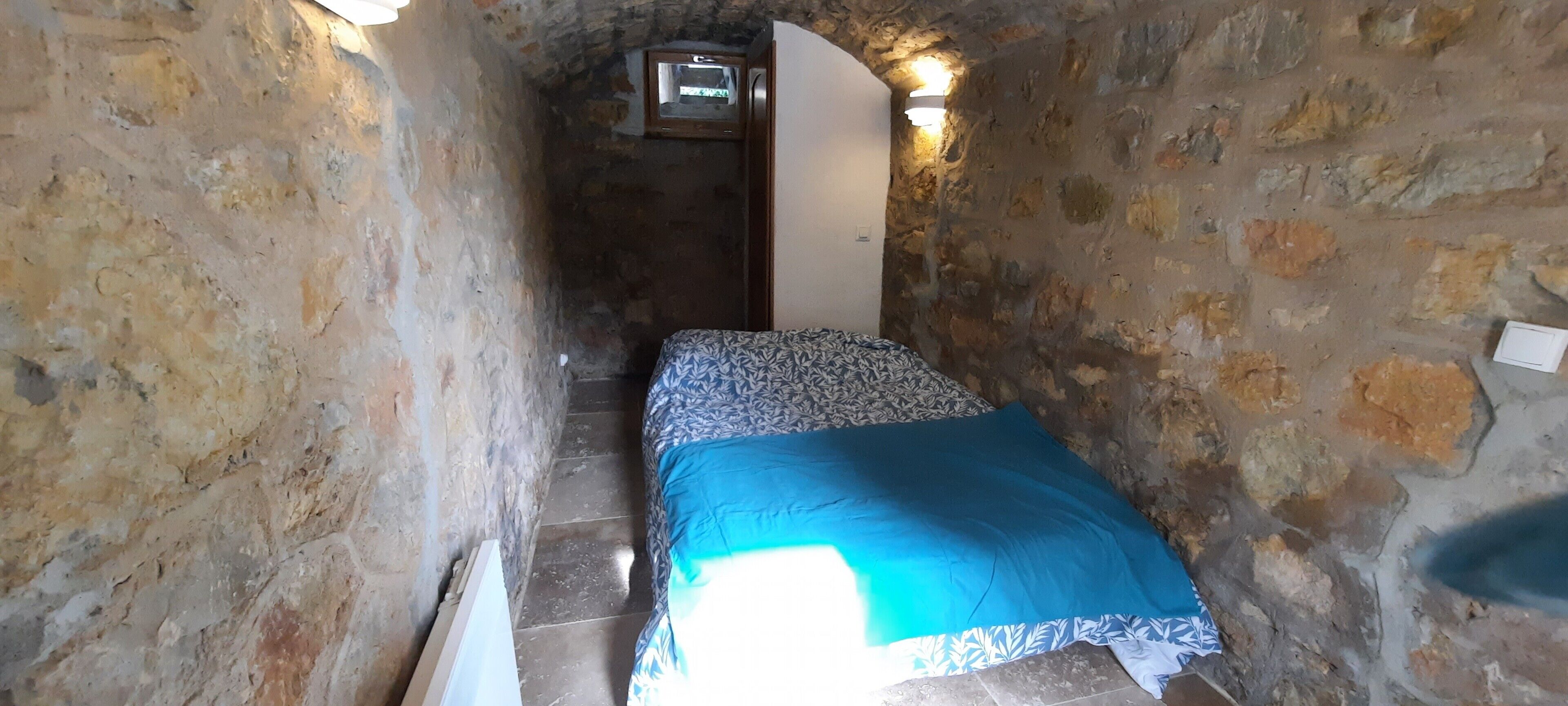A narrow stone-walled corridor leading to a bed with a patterned bedspread.