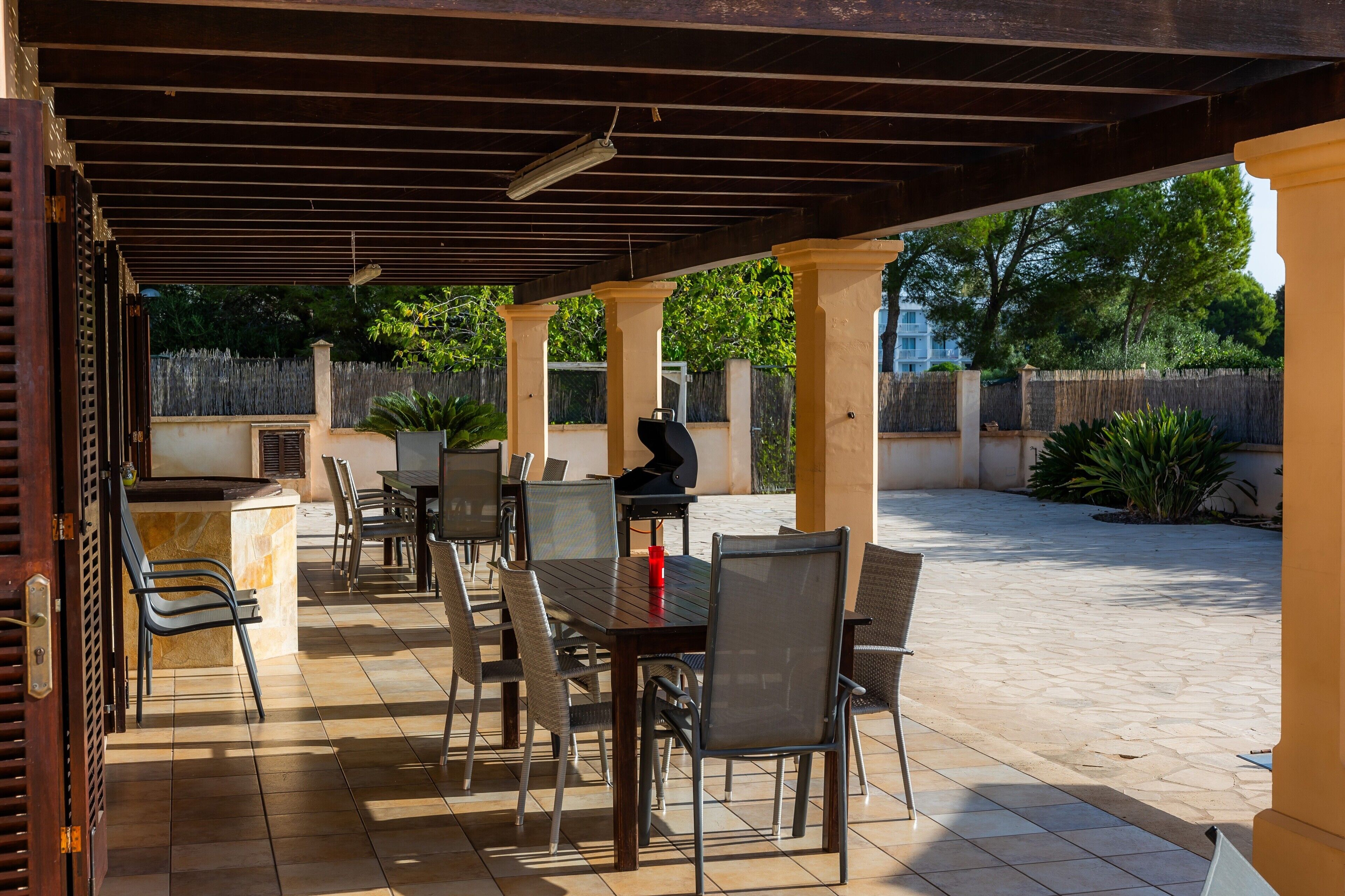 An outdoor patio with tables and chairs, a grill, and a view of a fenced area with trees.