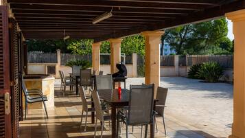 An outdoor patio with tables and chairs, a grill, and a view of a fenced area with trees.