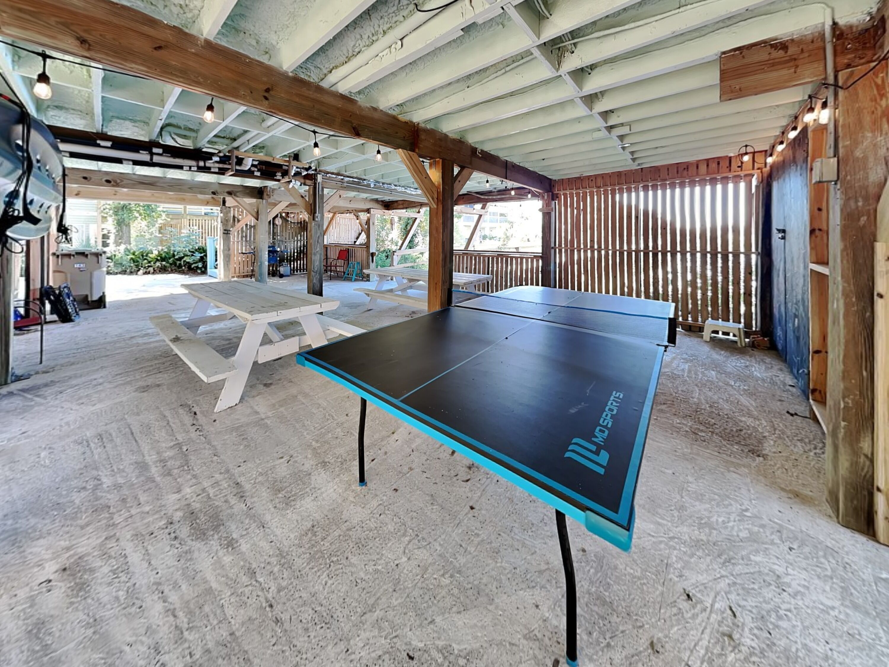 A ping pong table in a covered outdoor area with picnic tables and a wooden fence.