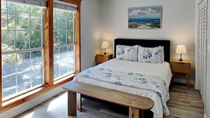 A bedroom with a large window, a bed with floral bedding, two bedside tables with lamps, a bench, and a ceiling fan. - Saltcreek Sunsets by Tybee Vacation Rentals (Tybee Island)