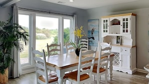 A dining area with a wooden table and white chairs, a white cabinet with glass doors, and a view of greenery through large windows.