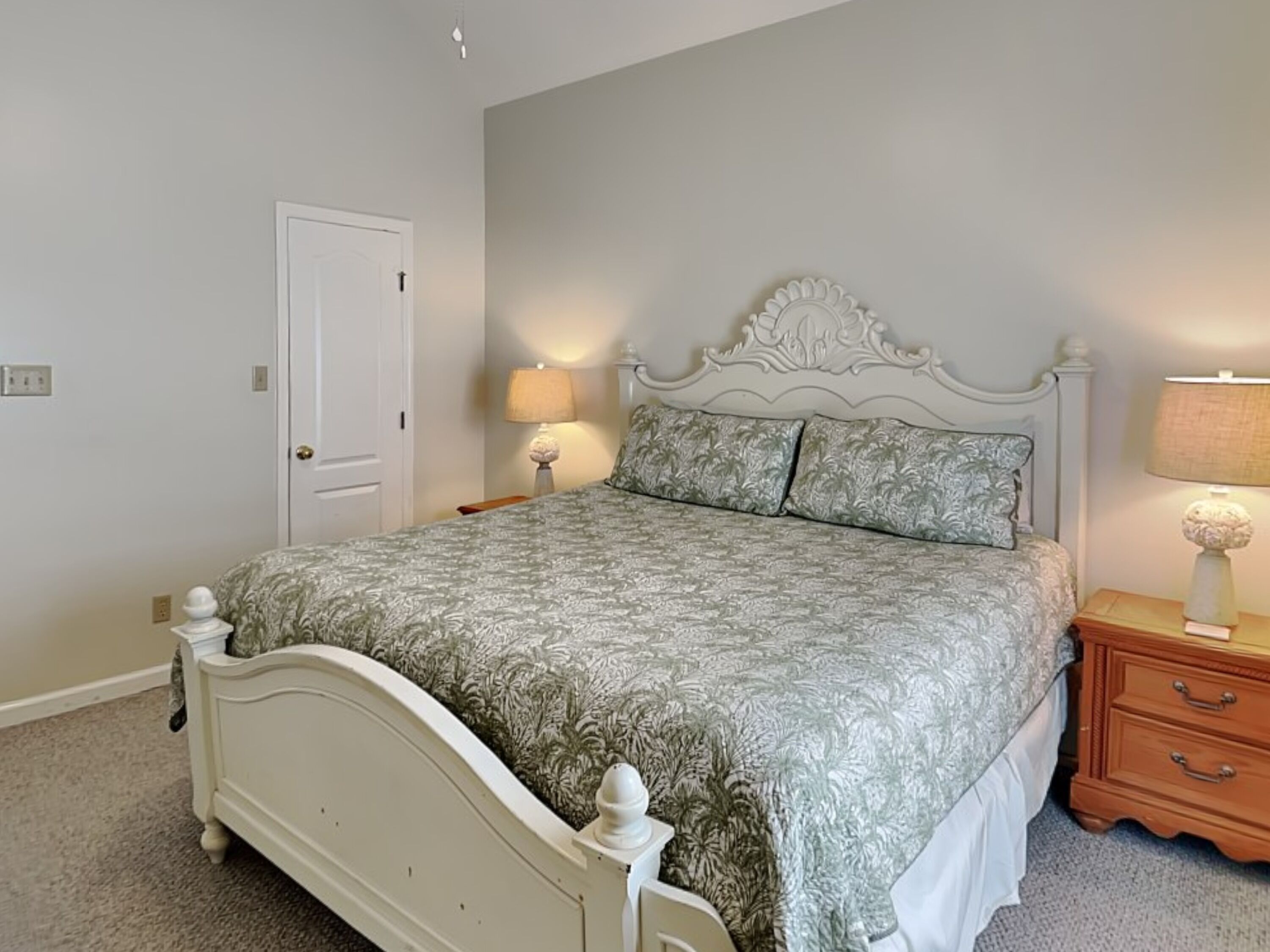 A bedroom with a white bed, floral patterned bedspread, two bedside lamps, a wooden nightstand, and a white door.