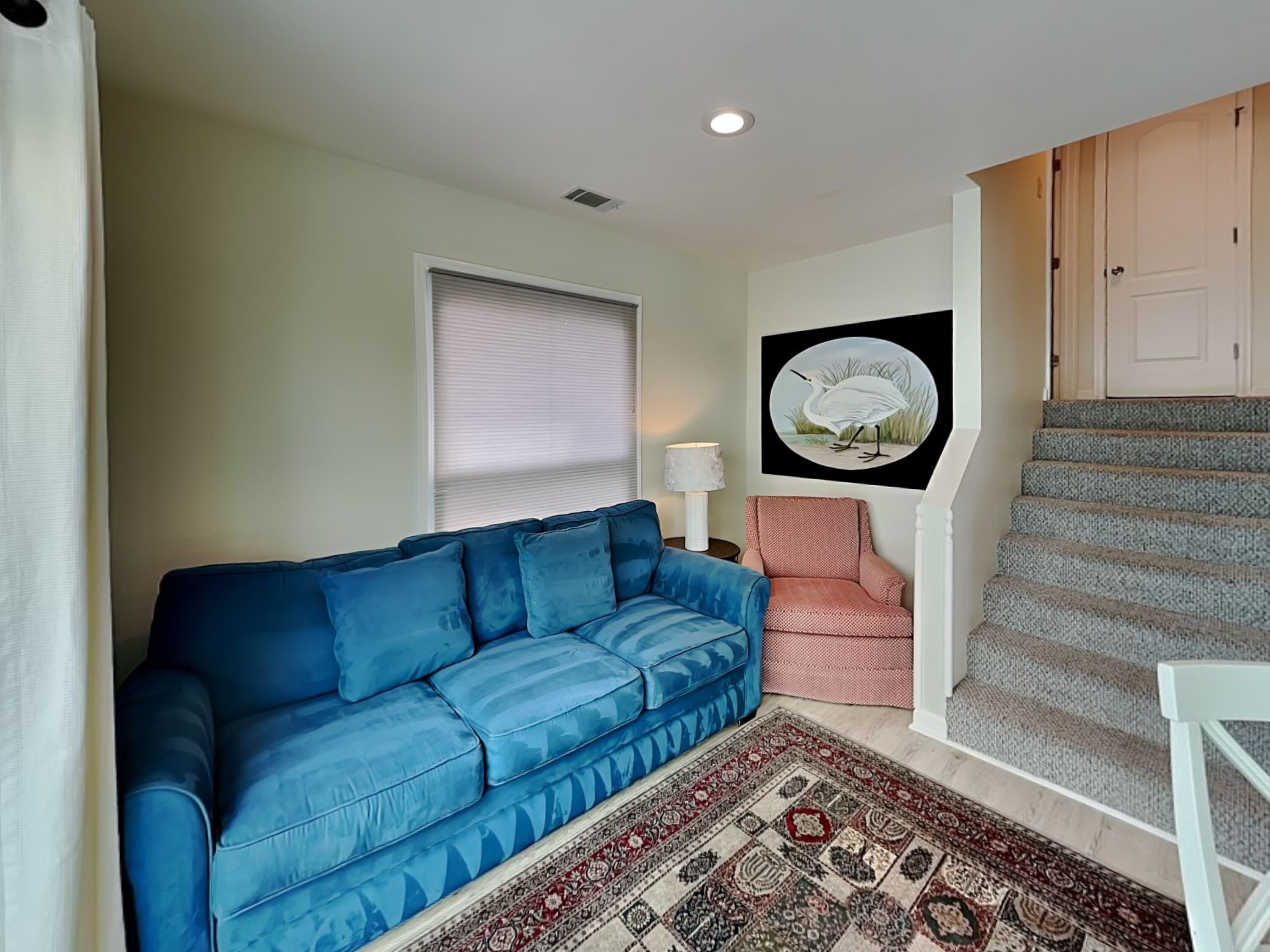 A living room with a blue sofa, a patterned rug, a pink chair, a lamp, and a staircase.