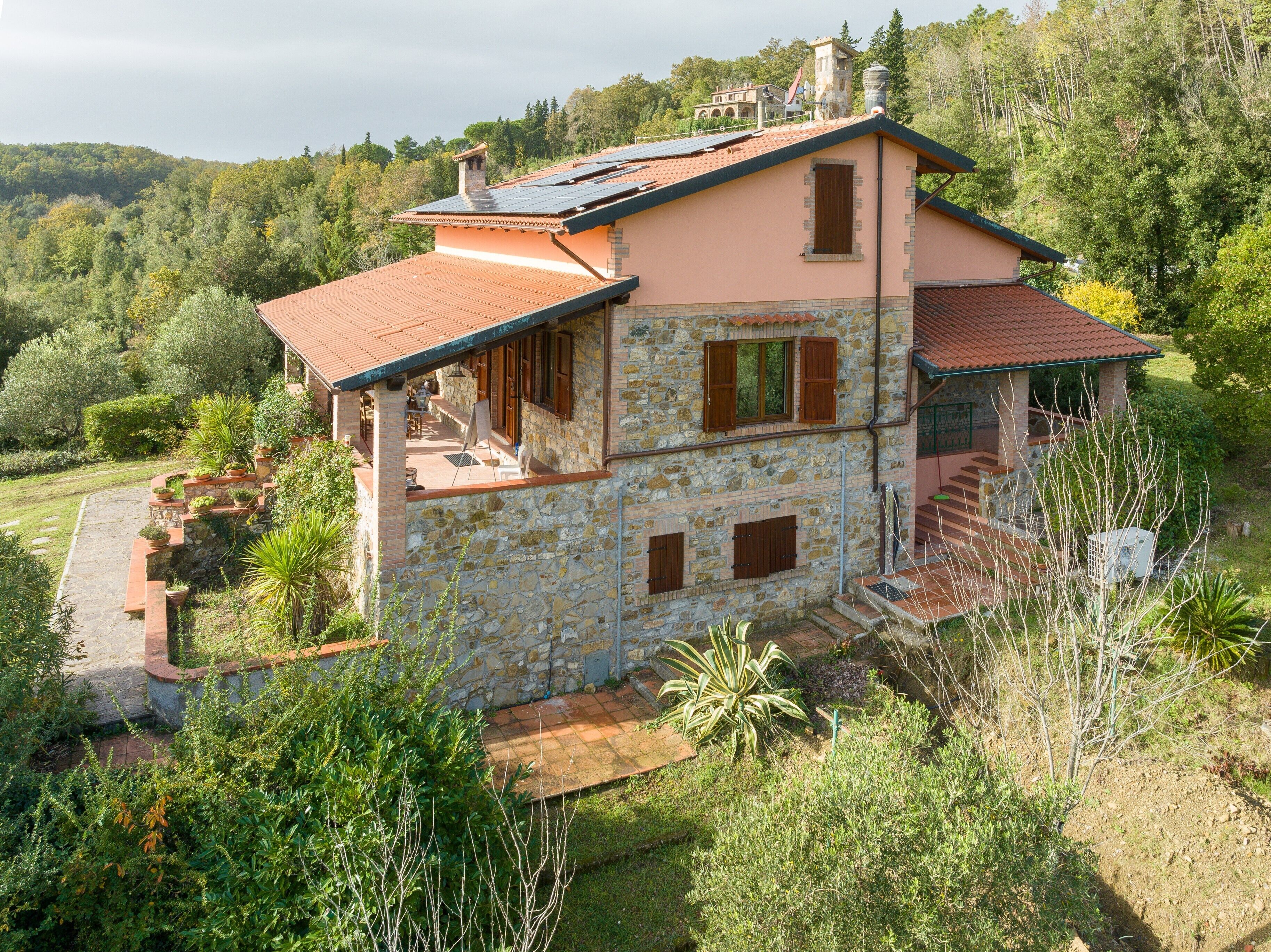 A two-story stone house with a red-tiled roof, a balcony, and a garden with various plants.