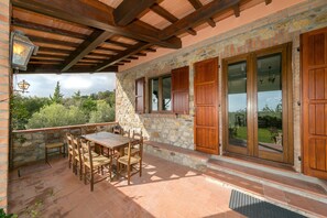 Un patio con una mesa y sillas, paredes de piedra, contraventanas de madera y vistas del paisaje circundante.