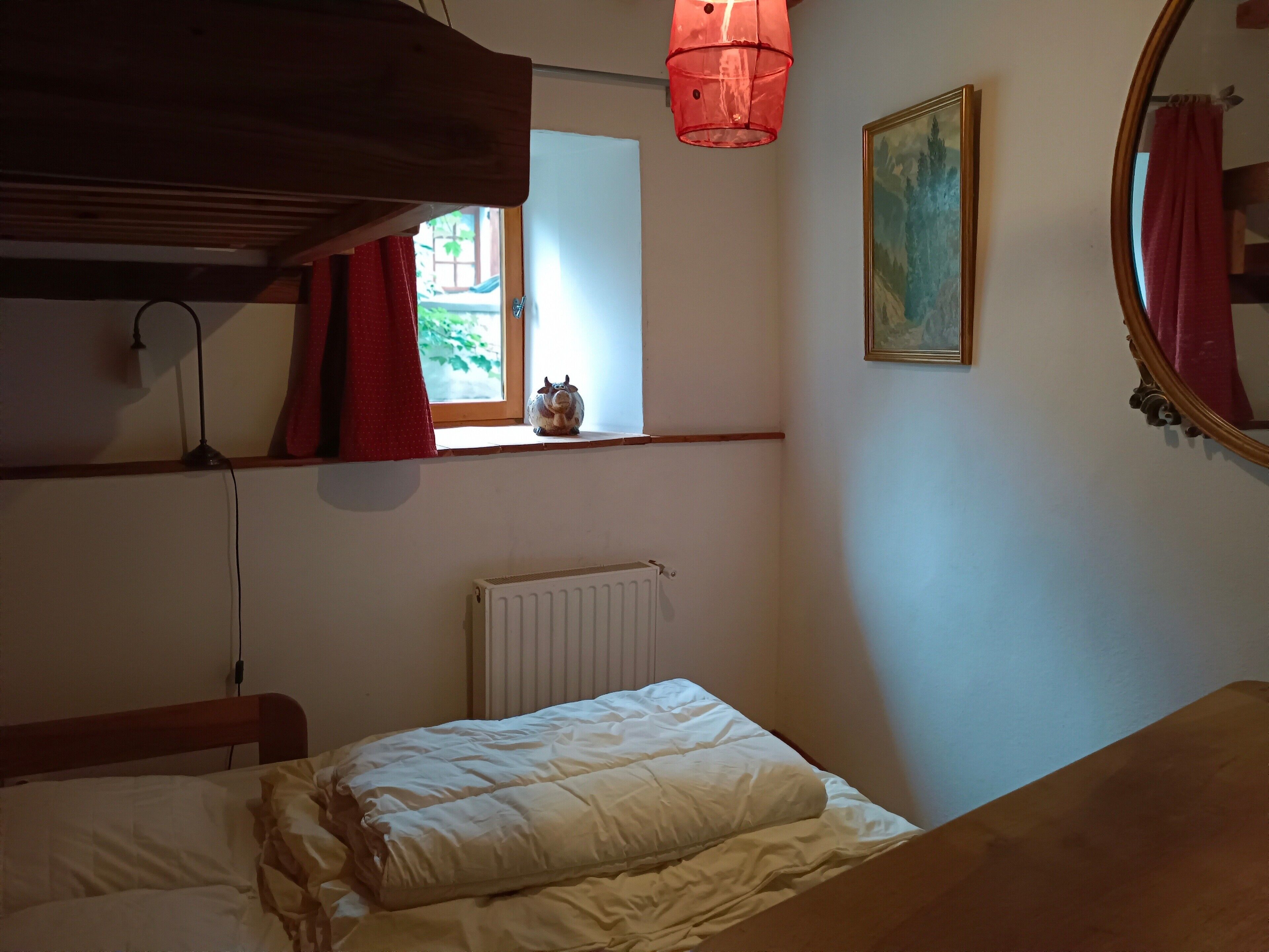 Um quarto com cama, radiador, janela com cortinas, uma pequena estante com uma figurinha e um quadro na parede.