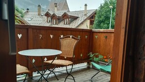 Een houten terras met een tafel en stoelen, een houten omheining met hartvormige uitsparingen, en een uitzicht op gebouwen en bergen op de achtergrond.