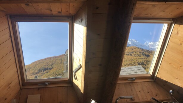 Een kamer met houten wanden en twee dakramen met uitzicht op een bebost berglandschap.
