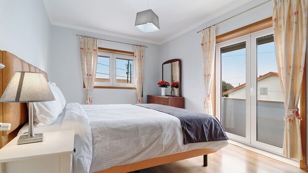 Un dormitorio con cama, una mesita de noche con lámpara, cómoda, espejo y ventana con cortinas.