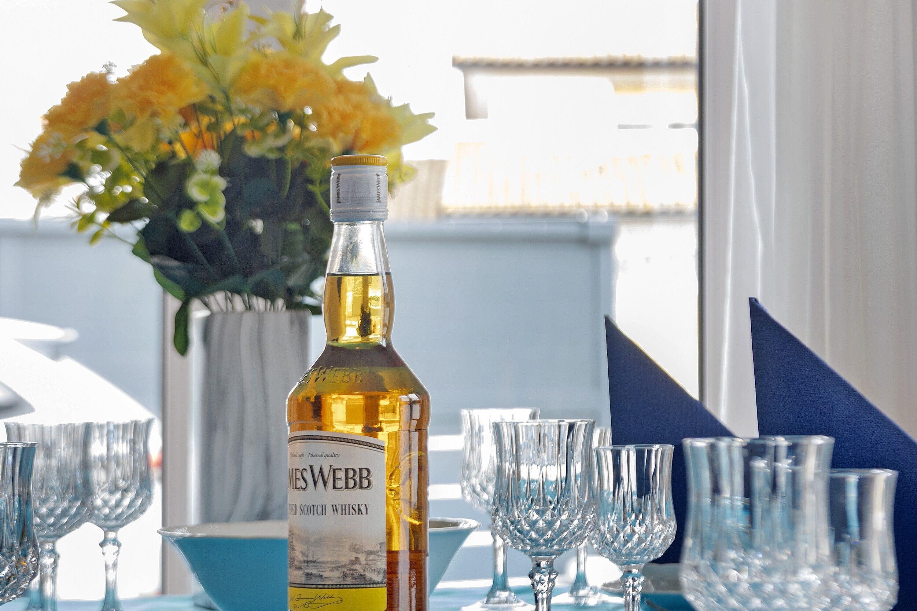 A bottle of whisky, several glasses, and a vase of flowers on a table.