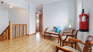 Un pasillo con suelo de madera, una escalera, una zona de estar con sillas y un banco, y un extintor de incendios montado en la pared.