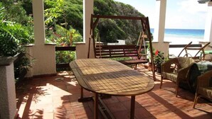 Eine überdachte Terrasse mit Tisch und Stühlen, umgeben von üppiger Vegetation und Blick auf den Ozean.