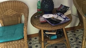 Ein Korbstuhl mit türkisblauer Sitzkissen, ein kleiner runder Tisch mit einer Lampe, eine Flasche und einige Zeitschriften.
