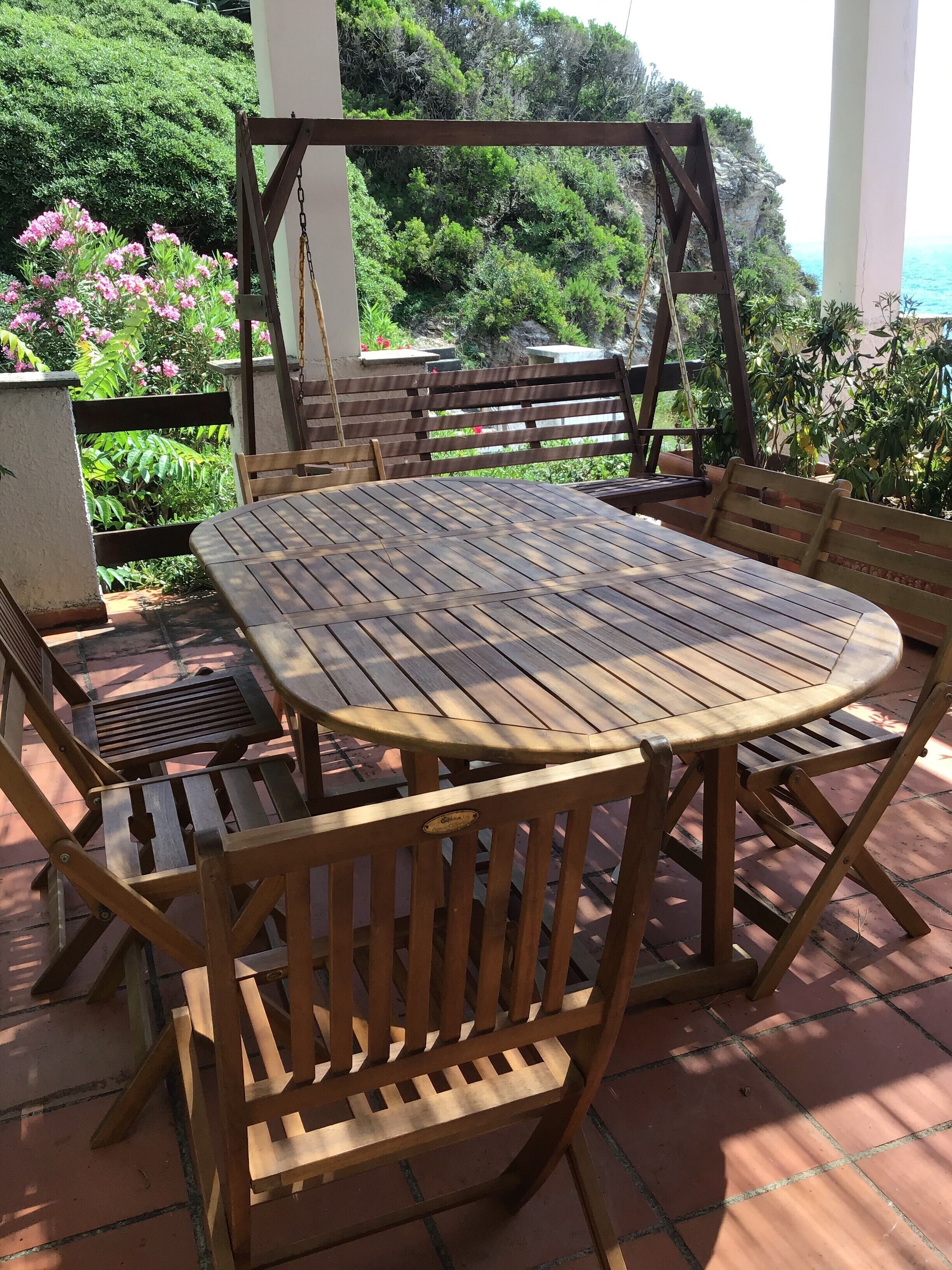 A wooden outdoor dining set with a swing chair, surrounded by lush greenery and a view of the sea.