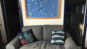 Ein graues Sofa mit zwei Zierkissen, darüber ein gerahmtes Kunstwerk mit Strandmotiv und eine Fernbedienung auf dem Sofa.