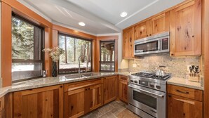 Une cuisine moderne dotée de meubles en bois, d’appareils électroménagers en acier inoxydable et de grandes fenêtres offrant une vue sur les arbres.