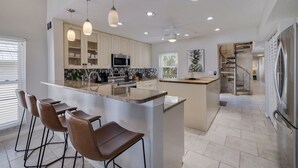 A modern kitchen with a central island, bar stools, pendant lights, and a stainless steel refrigerator. - 10 Pipers III – Beachfront Bliss with Ocean Views, Private Balcony & Grill Area (Indian Rocks Beach)