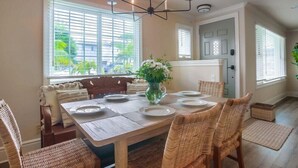 Un coin repas avec une table en bois dressée pour six personnes, entourée de chaises en osier, d’un lustre et de grandes fenêtres équipées de stores.