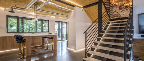 Un intérieur spacieux avec un escalier en bois, un coin cuisine doté d’un bar et de grandes fenêtres offrant une vue sur la verdure.
