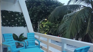 Une terrasse avec des chaises bleues et une table, entourée d’une balustrade blanche, d’où l’on peut admirer des palmiers et un mur orné de plantes grimpantes.
