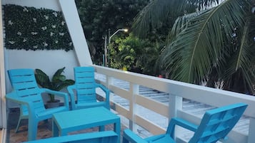 A terrace with blue chairs and a table, surrounded by a white railing, with a view of palm trees and a wall decorated with greenery.