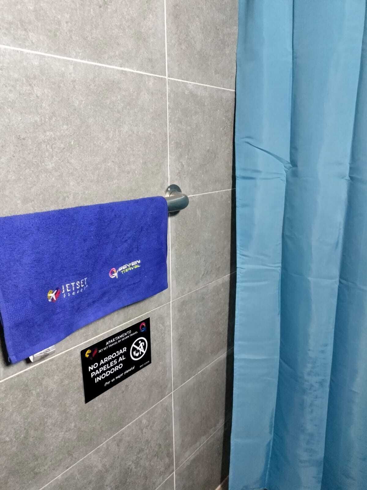 A bathroom with a blue towel hanging on a metal towel rack, a sign prohibiting paper towels, and a blue shower curtain.