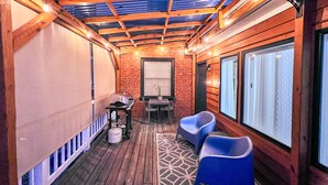 Un espace extérieur abrité, doté d’une terrasse en bois, d’un barbecue, de deux chaises bleues et d’un tapis à motifs.