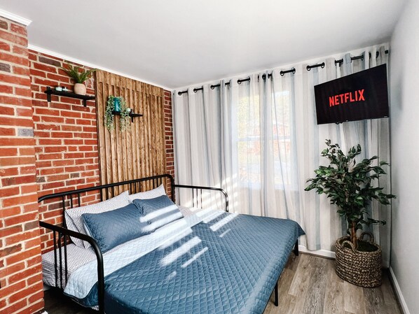 A bedroom with a brick wall, a wooden slat wall, a bed with a blue quilt, a potted plant, and a wall-mounted TV displaying 'NETFLIX'.