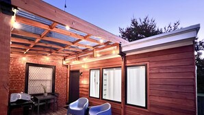 Une terrasse extérieure couverte, avec des murs en bois, deux chaises bleues, une table et des guirlandes lumineuses.