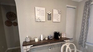 Une cuisine avec un plan de travail en bois, une machine à café et un vase rempli de fleurs. Deux éléments décoratifs muraux avec du texte sont présents, ainsi qu’une fenêtre avec des stores.
