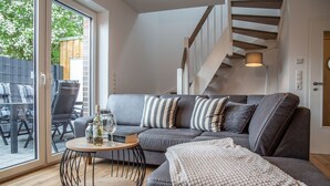 A modern living room with a grey sofa, striped pillows, a round wooden coffee table, and a staircase with wooden beams.
