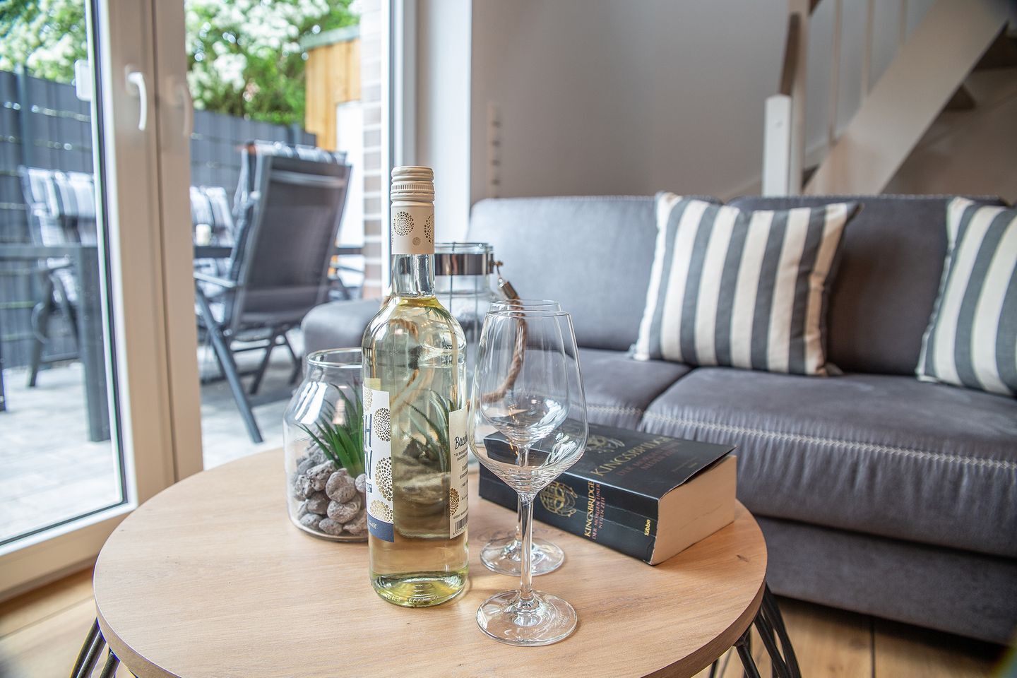 Une table ronde en bois, sur laquelle se trouvent une bouteille de vin blanc, deux verres à vin et un bocal décoratif rempli de petits cailloux. En arrière-plan, on aperçoit un canapé gris orné de coussins à rayures et un livre intitulé « Kings of the Hill ».