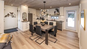 A modern kitchen with white cabinets, a central island, and a dining area with a wooden table and chairs. - La Châle-heureuse (Baie-Saint-Paul)