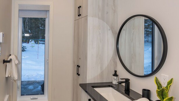 A modern bathroom with a wooden vanity, a round mirror, and a view of a snowy landscape through a window. - La Châle-heureuse (Baie-Saint-Paul)