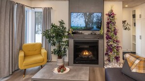 A modern living room with a fireplace, a television, a yellow armchair, a grey sofa, and a decorative plant. - La Châle-heureuse (Baie-Saint-Paul)