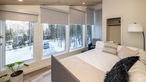 A bedroom with a large bed, a bedside table, a chair, a floor lamp, and a view of a snowy forest through large windows. - La Châle-heureuse (Baie-Saint-Paul)