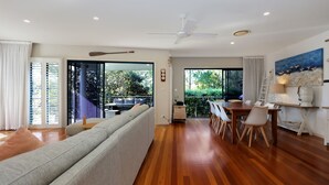 A modern living room with a sofa, dining table, and chairs. There is a balcony with a view of greenery. - 2 The Pier - Elegance by the Water (Tea Gardens)