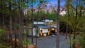 Une maison moderne avec une allée en gravier, entourée d’arbres, dotée de grandes fenêtres et d’un porche abrité.