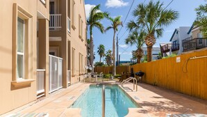 A swimming pool with a wooden fence, palm trees, and a building with balconies. - Cozy Coastal Escape w/ Pool - 1/2 Block to Beach! (South Padre Island)