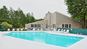 A rectangular swimming pool with clear blue water, surrounded by white lounge chairs and umbrellas, with a building featuring a flat roof and large windows in the background.