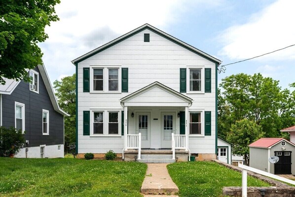 A two-story white house with green shutters, a front porch, and a small attached garage.