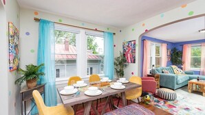 Ein Essbereich mit einem Tisch für vier Personen, Stühlen und einer Bank. Zwei Fenster mit Vorhängen, eine Couch und ein kleiner Tisch mit einer Pflanze sind ebenfalls vorhanden.