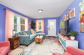 Una sala de estar con un sofá azul, una mesa de centro de madera y una silla rosa. Hay dos ventanas con cortinas rosas, una puerta blanca y un televisor sobre un mueble blanco.