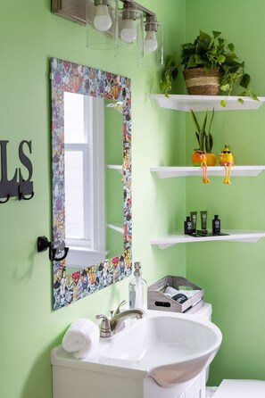 Bany amb una paret verda, un lavabo blanc, un mirall amb una estructura de colors, i prestatgeries amb plantes i productes de tocador.
